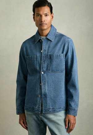 Jeansjacke in Mittelblau, mit Knopfleiste, zwei großen Brusttaschen und klassischem Kragen. Leichte Textur, lässige Passform.