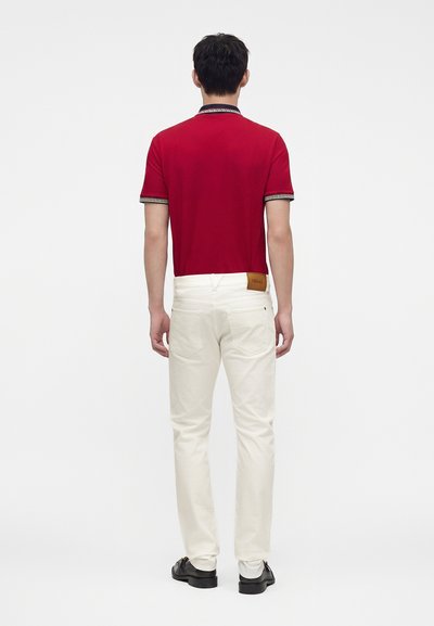Homme debout, tourné de dos, portant un polo rouge à manches courtes, un pantalon blanc avec un écusson en cuir de marque, et des chaussures noires sur un fond uni.