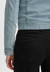 Chaqueta de mezclilla azul claro con costuras detalladas, puño abotonado, combinada con jeans negros que tienen un bolsillo trasero. Enfoque en el material y el diseño.