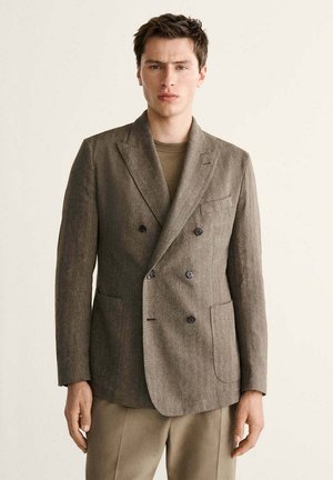 Junger Mann mit braunem, doppelt geknöpftem Blazer und passenden Hosen, der vor einem schlichten hellen Hintergrund steht.