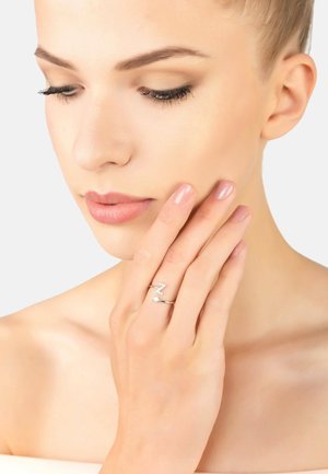 Silver ring with a zigzag design, featuring a small gemstone accent. Model's hand is gently touching her face, highlighting the ring.