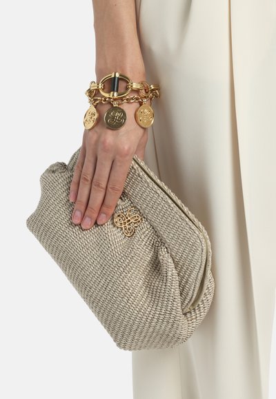 Main tenant une pochette tissée beige ornée d'un embleme en or, portant un bracelet à charms en or avec trois pendentifs ronds en forme de pièce.