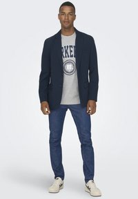 Marineblazer über einem grauen Grafik-T-Shirt, kombiniert mit dunklen Jeans und weißen Sneakers. Glatter Stoff, taillierte Passform und minimale Details.
