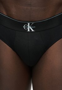Μαύρα εσώρουχα Calvin Klein φορεμένα σε σκούρο δέρμα, με ζώνη που δείχνει το λευκό λογότυπο "cK calvin klein".