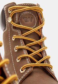 Botas de cuero marrón con ojales hexagonales, cordones amarillos y marrones, costuras blancas y un logo circular en la lengüeta.