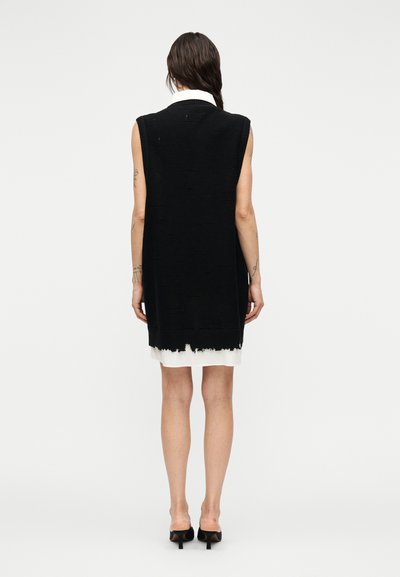 Μαύρο πλεκτό φόρεμα χωρίς μανίκια με εμφανή ξεφτισμένα τελειώματα και υφή στην επιφάνεια· λευκή υποφούστα φαίνεται να ξεπροβάλλει από κάτω.