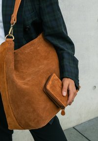 Brown suede shoulder bag with a soft texture and curved shape, accompanied by a matching wallet. The bag features a fabric shoulder strap.