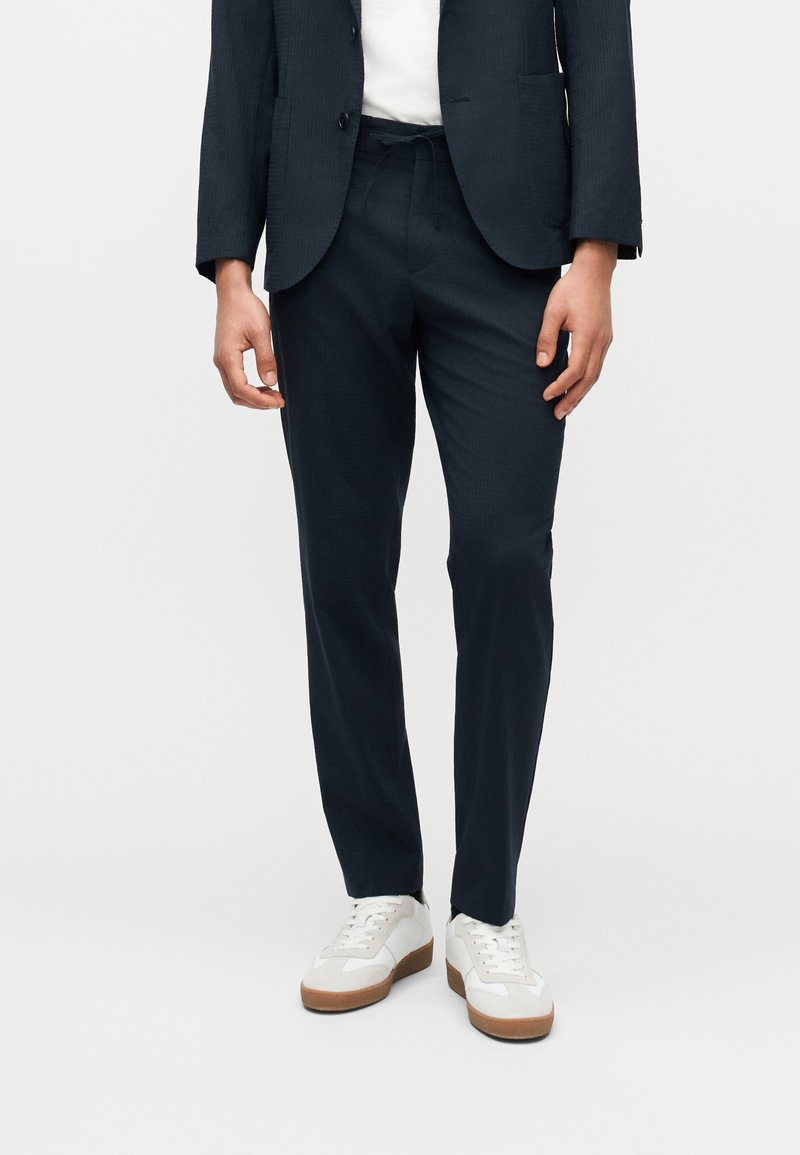Homme portant un pantalon sur mesure bleu marine foncé, un blazer assorti, une chemise blanche et des baskets blanches à semelles marron, se tenant devant un fond uni.