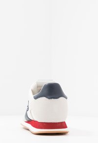 Sneaker bianco con dettagli in pelle blu navy, una finitura in suede rosso sulla suola, superfici testurizzate e un design del tallone curvo.