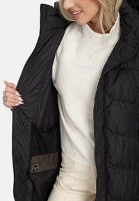 Naine kannab musta polsterdatud jope valge sviitri peal, hoides jope lahti, et näidata sees olevat võrgupocketit, kus on nutitelefon.