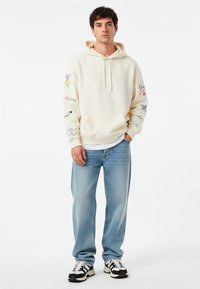 Felpa cremosa con design ricamati in modelli colorati. Tasca frontale a marsupio, polsini a costine, abbinata a jeans azzurri chiari e sneakers nere.