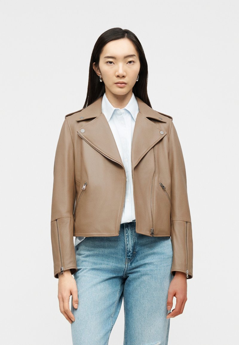 Γυναίκα που φοράει μια καφέ δερμάτινη μπουφάν μοτοσικλετιστή πάνω από ένα λευκό πουκάμισο με γιακά και ανοιχτό μπλε τζιν, με φόντο έναν απλό φωτεινό τοίχο.