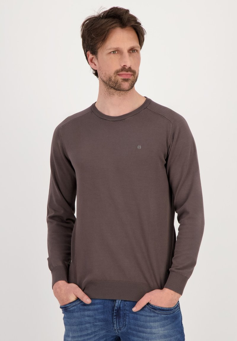 Maglione marrone lavorato a maglia con maniche lunghe, colletto rotondo, polsini e orlo a coste. Dettaglio del logo in piccolo sul petto. Abbinato a jeans blu.