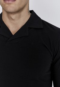 Camisa polo negra de manga larga, confeccionada en un material suave, con un cuello plano y el logo en relieve "Siksilk" en el lado superior izquierdo.