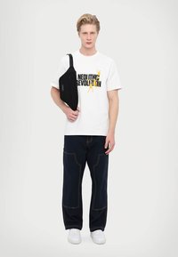 Tricou alb cu grafica neagră și galbenă, geantă de umăr neagră, pantaloni negri largi cu cusături contrastante și adidași albi.
