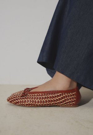 Chaussure plate tissée rouge et beige avec un petit nœud portée avec un pantalon bleu foncé sur un fond de sol neutre.