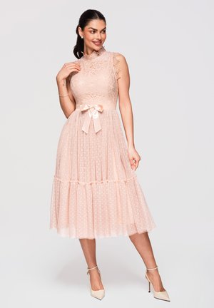 Mujer con vestido midi rosa de encaje sin mangas con cinturón de lazo de satén, falda plisada y zapatos de tacón beige, de pie y sonriendo.