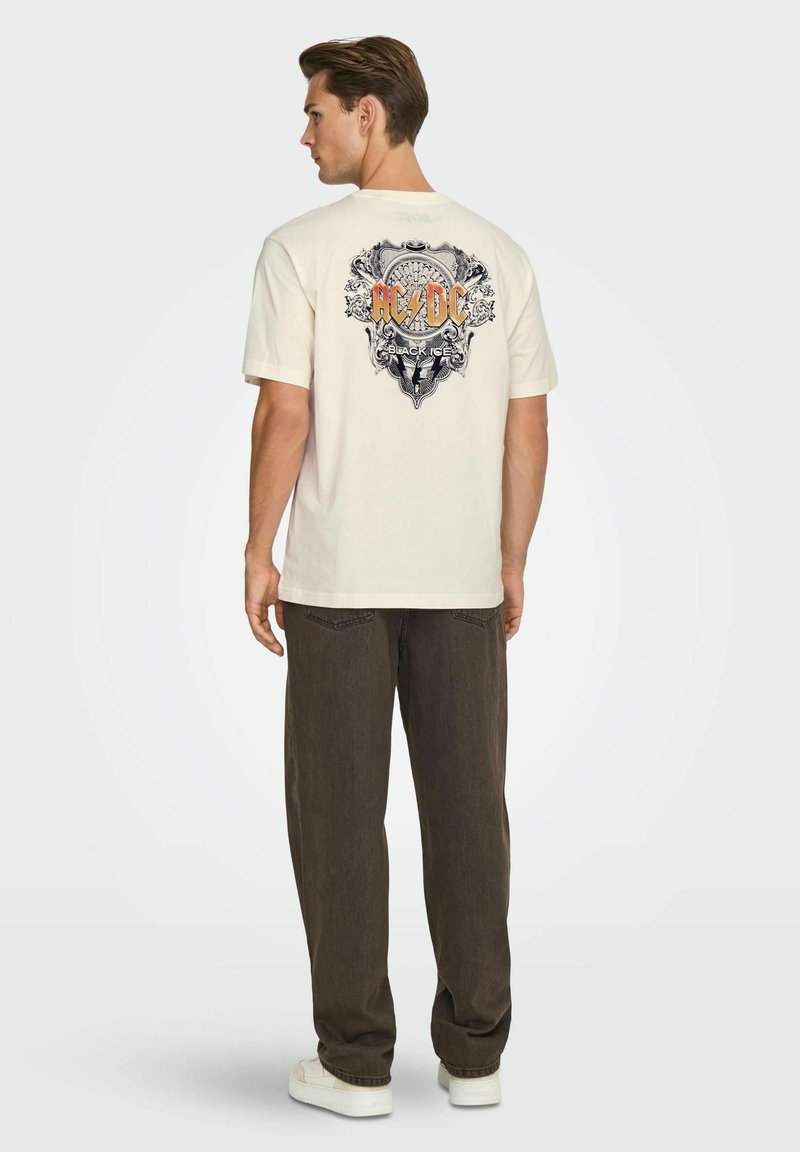 Cream-colored cotton t-shirt featuring a detailed AC/DC graphic on the back, paired with dark brown pants and white sneakers.