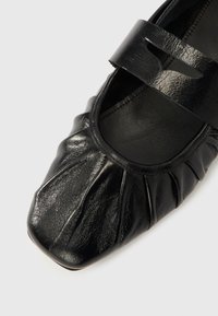 Czarny skórzany but o zaokrąglonym czubku, z marszczoną teksturą na cholewce i paskiem na podbiciu z wyciętym wzorem.