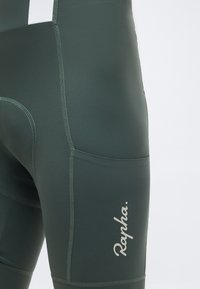 Κοντινό πλάνο σκούρων πράσινων σορτς ποδηλασίας με εμφανείς ραφές και λευκό λογότυπο «Rapha» στην περιοχή της πλαϊνής τσέπης.