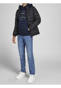 Svart pufferjacka med dragkedja, marinblå hoodie under med grafisk text, ljusblå jeans och vita sneakers.