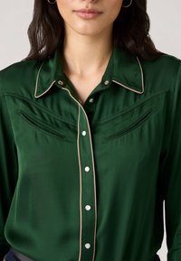 Grüne Satinbluse mit spitzem Kragen, Perlenknöpfen und kontrastierender beiger Einfassung. Enthält zwei dekorative Fronttaschen.