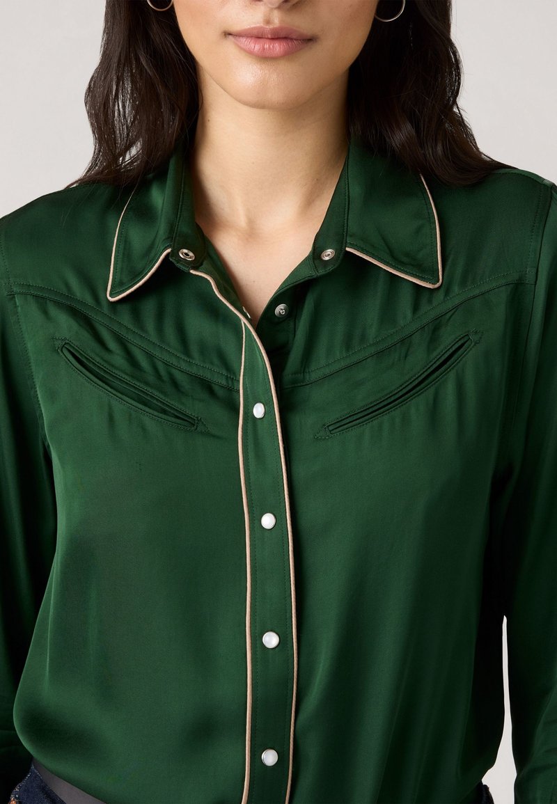 Green satin blouse featuring a pointed collar, pearl buttons, and contrasting beige piping. Includes two decorative front pockets.