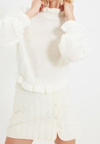 Biały sweter dzianinowy z falbaną przy kołnierzu i mankietach, w połączeniu z pikowaną spódnicą w kolorze off-white z listwą guzikową.