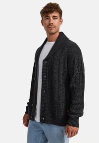 Cárdigan de punto gris oscuro con textura de canalé, gran cuello de chal y cierre de botones, combinado con una camiseta blanca y jeans claros.