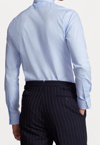 Světle modrá košile s dlouhými rukávy, manžety na knoflíky; nošená s tmavými kalhotami s drobnými pruhy. Hladká látka, střih na míru, minimalistický design.
