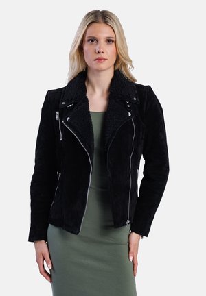 Een vrouw met lang blond haar draagt een zwart suède jack met een schapenvacht kraag over een groene jurk, staande tegen een witte achtergrond.