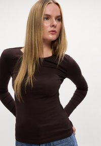 Hnedý dlhý rukáv vyrobený z elastického materiálu, s okrúhlym výstrihom a slim fit strihom. Skombinované s neformálnymi modrými džínsami.