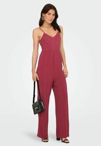 Combinaison bordeaux à fines bretelles, décolleté en V et jambes larges ; associée à un sac en bandoulière noir et des talons noirs à lanières.