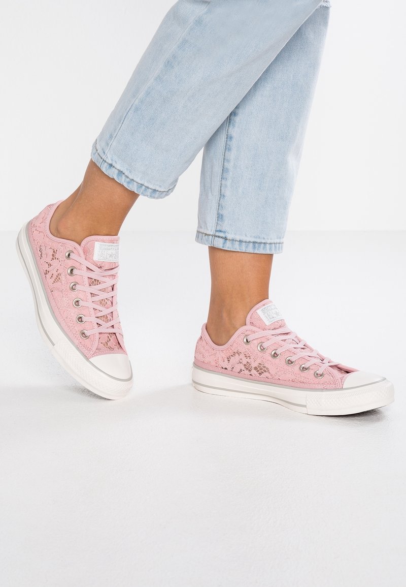 Pieds portant un jean bleu clair et des baskets basses roses en dentelle avec semelles et lacets blancs sur fond blanc.