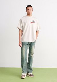 T-shirt oversized bege claro com um logotipo vermelho e preto, combinado com jeans reto verde desbotado e sapatilhas castanhas.