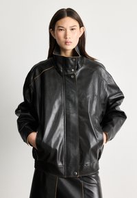 Crna kožna bomber jakna s visokim ovratnikom, s kontrastnim narančastim šavovima, drukerima i širokim krojem. Glatka tekstura.