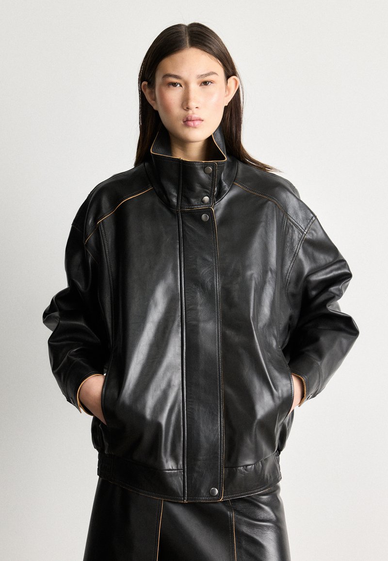 Crna kožna bomber jakna s visokim ovratnikom, s kontrastnim narančastim šavovima, drukerima i širokim krojem. Glatka tekstura.