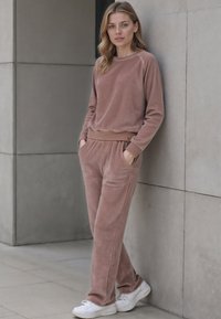 Měkká loungewear souprava v jemné růžové barvě vyrobená z hebké látky, obsahující top s kulatým výstřihem a kalhoty s elastickým pasem a kapsami, doplněná bílými teniskami.
