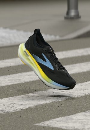 Černo-modrá běžecká bota Brooks se žlutou mezipodešví plující nad městským přechodem na asfaltu.