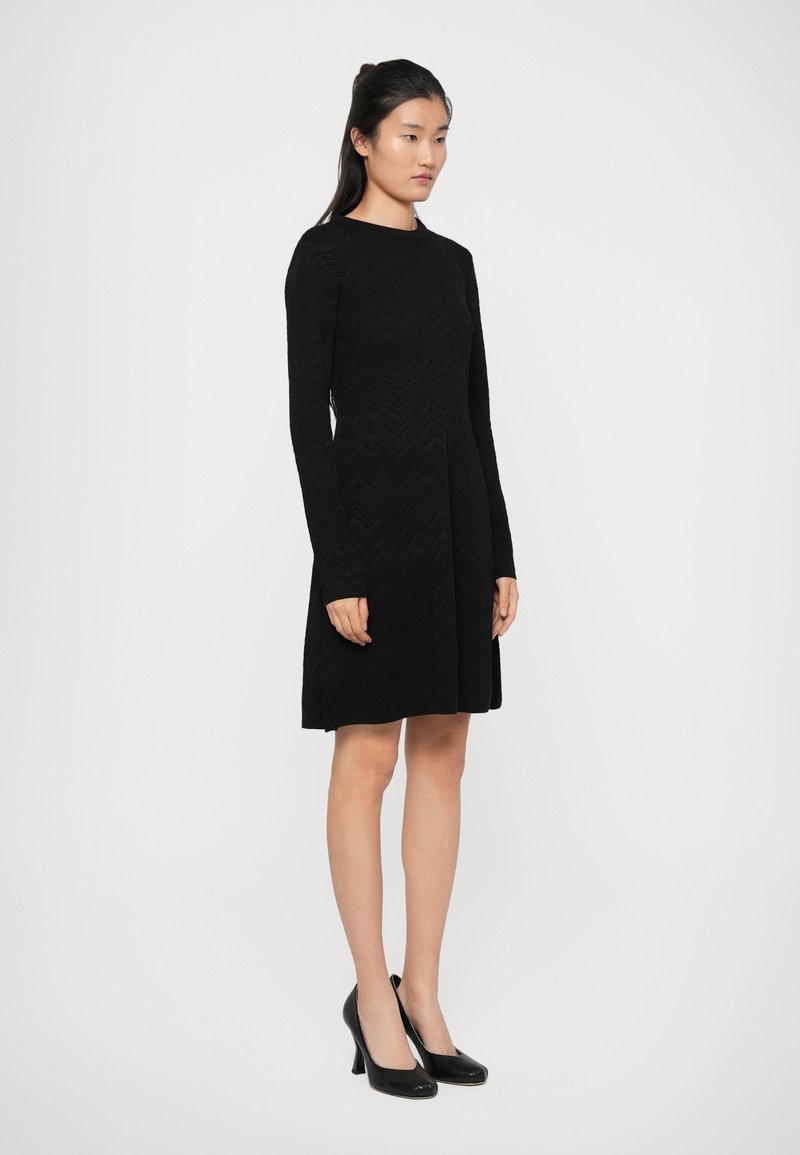 Robe noire à manches longues avec un motif en chevron texturé, jupe à mi-genoux et corsage ajusté, portée avec des chaussures à talons hauts noires.