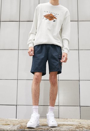 Person trägt weißen Air Jordan Pullover, dunkelblaue Shorts, weiße Socken und weiße Nike-Sneaker und steht vor einer gefliesten Wand.
