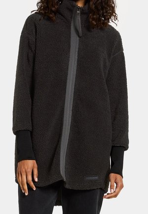 Person trägt eine lange schwarze Fleecejacke mit Frontreißverschluss und Daumenlöchern an den Ärmeln, steht vor weißem Hintergrund.