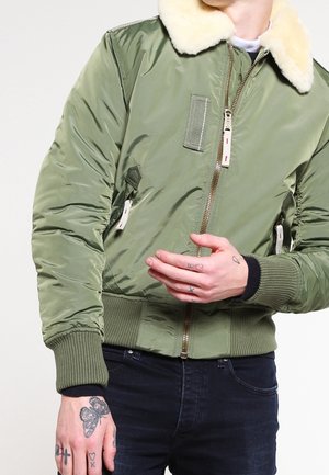 Zielona kurtka bomberka z kremowym kołnierzem z owczej skóry, zamkiem błyskawicznym z przodu i ściągaczami na mankietach, noszona przez osobę z licznymi tatuażami na rękach.