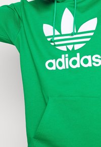 Zelená mikina s kapucí vyrobená z bavlny, s velkým bílým logem Adidas a klasickým designem se třemi lístky. Obsahuje nastavitelná stahovací šňůrky a přední kapsu.