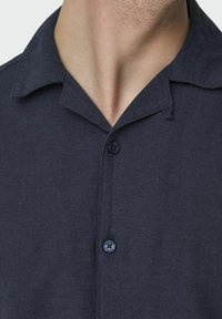 Camisa de botones azul marino oscura fabricada en un tejido ligero, con cuello y un cierre de un solo botón, que presenta un acabado texturizado.