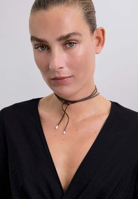 Brązowy skórzany naszyjnik z regulowanym zapięciem, z końcówkami w kolorze srebrnym. Modelka ma na sobie czarną bluzkę z dekoltem w serek. Gładka tekstura, prosty design.