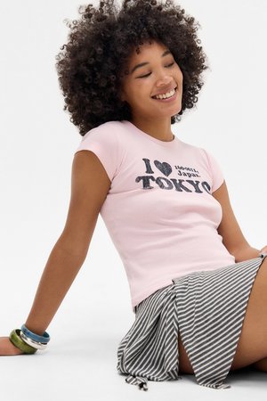 Camiseta ajustada de color rosa claro con gráfico de "I ♥ Tokyo" en impresión oscura. Combinada con una falda a rayas gris. Los accesorios incluyen pulseras coloridas.