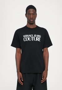 Juoda medvilninė marškinėliai su ryškiu baltu užrašu "VERSACE JEANS COUTURE." Standartinio kirpimo, trumpomis rankovėmis, apvali iškirptė. Paprastas dizainas.