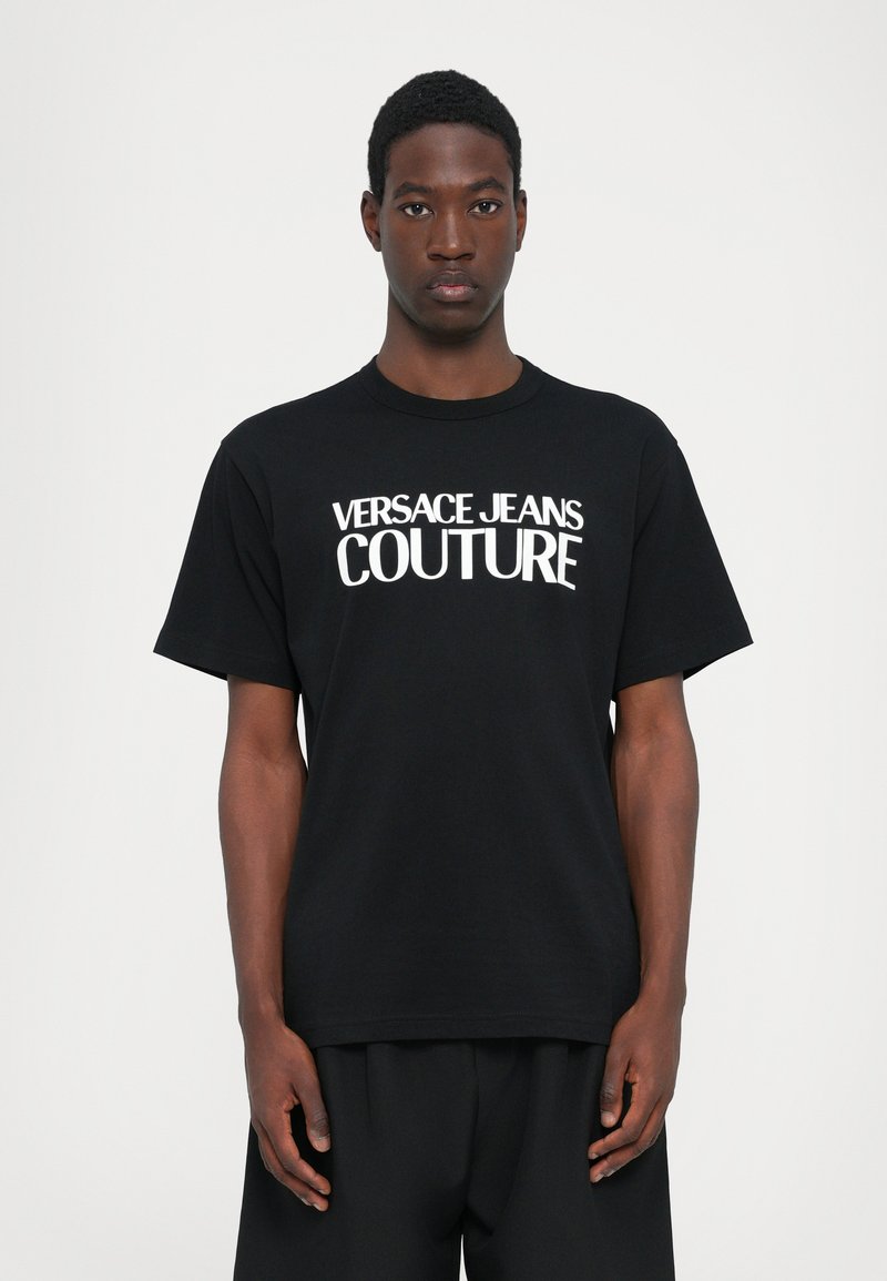 Juoda medvilninė marškinėliai su ryškiu baltu užrašu "VERSACE JEANS COUTURE." Standartinio kirpimo, trumpomis rankovėmis, apvali iškirptė. Paprastas dizainas.