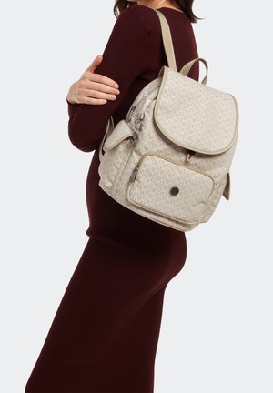 Sac à dos beige à motif avec fermeture à rabat, bretelles réglables, poche avant et ferrures argentées, porté sur une robe bordeaux.